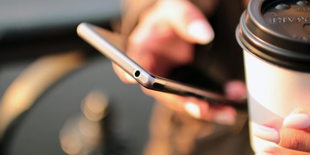 Person holding a smartphone and a takeaway coffee cup while texting outdoors.